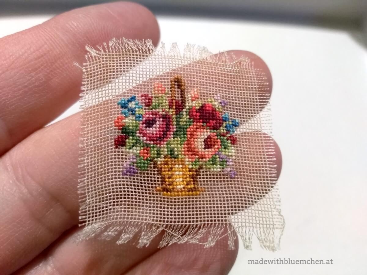 Eine Miniaturstickerei, die einen Blumenkorb zeigt, liegt auf drei Fingerspitzen. 
